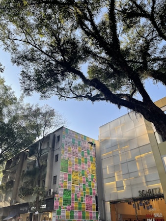 A street scene featuring a tall building covered with colorful posters advertising various music genres such as rock, pop, and funk. The posters are arranged in a grid pattern with vibrant colors including pink, green, and yellow. Adjacent to this building is a modern structure with frosted glass panels. In the foreground, large trees with lush green foliage are visible, casting shadows in the bright daylight.