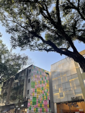 A street scene featuring a tall building covered with colorful posters advertising various music genres such as rock, pop, and funk. The posters are arranged in a grid pattern with vibrant colors including pink, green, and yellow. Adjacent to this building is a modern structure with frosted glass panels. In the foreground, large trees with lush green foliage are visible, casting shadows in the bright daylight.