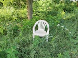 A white plastic chair is placed amidst lush green foliage and tall grass, highlighting an overgrown outdoor setting. A tree trunk is visible in the background, partially shaded by the surrounding greenery.