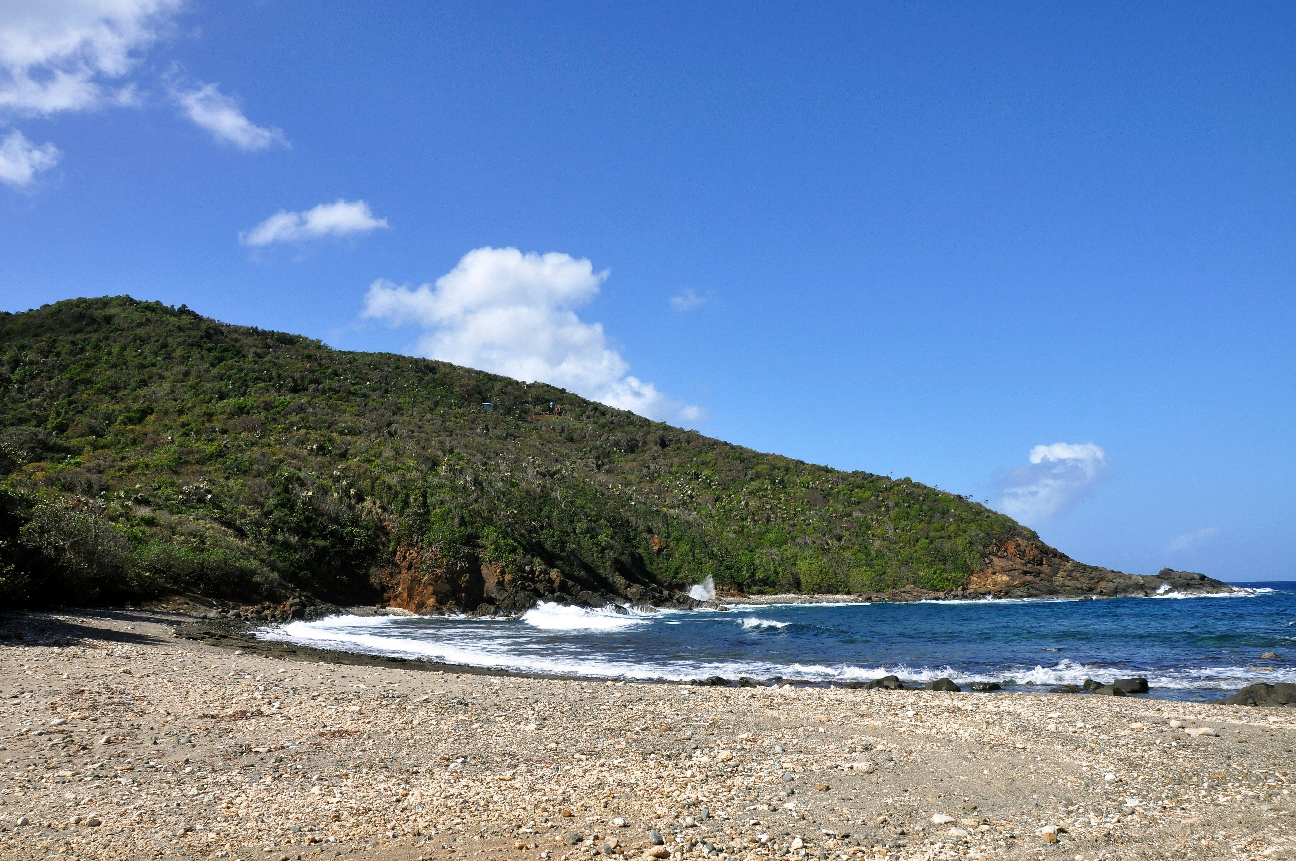 A blue, puffy white cloud view of a natural tropical rock and coral rubble sand beach and waves with green hillside descending to the water junction of the Caribbean Sea and Atlantic Ocean.