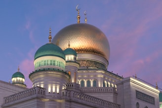 A mosque with a large gilded dome and several smaller green domes, adorned with intricate patterns and Arabic script. The architecture is illuminated by soft lighting against a twilight sky with shades of pink and purple.