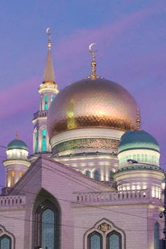 A stunning mosque with a large gold dome and multiple minarets, set against a vibrant twilight sky. The architectural details include intricate patterns and Arabic script on the domes, while the structure is illuminated with soft lighting.