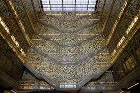 An architectural interior with multiple levels featuring ornate patterns on the walls and a large glass roof allowing natural light to flood in. The design creates an intricate and mesmerizing visual effect with a complex mosaic of light and shadow.