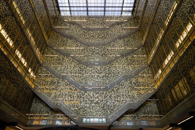 An architectural interior with multiple levels featuring ornate patterns on the walls and a large glass roof allowing natural light to flood in. The design creates an intricate and mesmerizing visual effect with a complex mosaic of light and shadow.