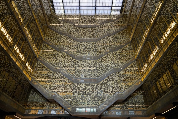 An architectural interior with multiple levels featuring ornate patterns on the walls and a large glass roof allowing natural light to flood in. The design creates an intricate and mesmerizing visual effect with a complex mosaic of light and shadow.