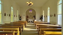 The interior of a simple, well-lit church with wooden pews arranged on either side of a central aisle leading to an altar. The walls are light-colored and lined with stained glass windows. Religious statues and plaques are mounted along the walls. A chandelier hangs from the ceiling, illuminating the space along with natural light streaming through the windows.