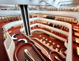An interior view of a multi-level library with a modern architectural design, featuring open floors and spiral staircases. Bookshelves line the walls, and there are multiple rows of tables and seating areas where people are engaged in reading and studying. The color scheme predominantly includes red carpeting and white structural elements.