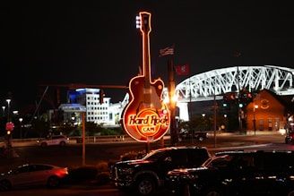 A brightly lit sign in the shape of a guitar is prominently displayed against a dark night sky. The sign reads 'Hard Rock Cafe' in glowing red and yellow lights. In the background, an illuminated bridge and several buildings are visible. Cars are parked below the sign, and there are a few flags nearby.
