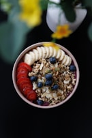 A bowl filled with a variety of healthy ingredients including sliced bananas, strawberries, blueberries, granola, and almond slices. The arrangement looks appetizing and carefully organized. There is a blurred flower in the background adding a touch of natural beauty.