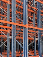A complex structure composed of multiple metal beams and girders, mainly in dark blue and orange hues. The intricate, crisscrossing framework suggests the interior of an industrial racking system, possibly used for storage or support.
