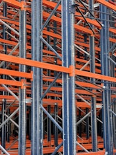 A complex structure composed of multiple metal beams and girders, mainly in dark blue and orange hues. The intricate, crisscrossing framework suggests the interior of an industrial racking system, possibly used for storage or support.