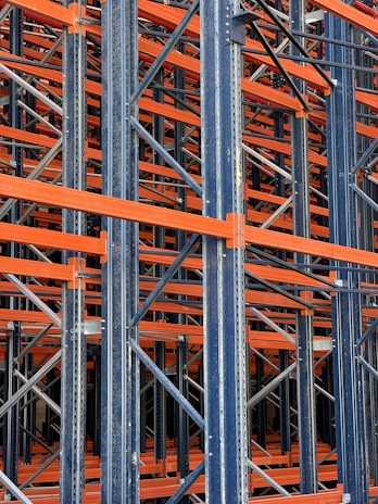 A complex structure composed of multiple metal beams and girders, mainly in dark blue and orange hues. The intricate, crisscrossing framework suggests the interior of an industrial racking system, possibly used for storage or support.