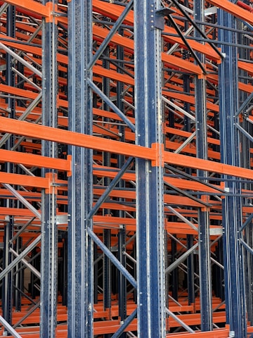 A complex structure composed of multiple metal beams and girders, mainly in dark blue and orange hues. The intricate, crisscrossing framework suggests the interior of an industrial racking system, possibly used for storage or support.