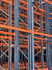 A complex structure composed of multiple metal beams and girders, mainly in dark blue and orange hues. The intricate, crisscrossing framework suggests the interior of an industrial racking system, possibly used for storage or support.