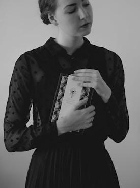 A woman is holding a book close to her chest with her eyes gently closed. She is wearing a black polka-dotted blouse and a dark skirt. The photograph is in black and white, creating a vintage and thoughtful atmosphere.