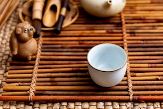 A small ceramic cup sits on a bamboo mat alongside a ceramic figurine resembling an animal. The arrangement includes tea utensils and a teapot, creating a serene and traditional tea setting.