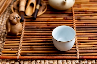 A small ceramic cup sits on a bamboo mat alongside a ceramic figurine resembling an animal. The arrangement includes tea utensils and a teapot, creating a serene and traditional tea setting.