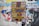 A vibrant poster with bold, colorful text and a zigzag lightning bolt design is pasted onto a textured brick wall. The surrounding area is layered with torn scraps of other posters and paper, creating a collage effect.