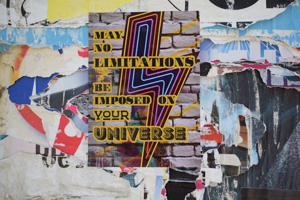 A vibrant poster with bold, colorful text and a zigzag lightning bolt design is pasted onto a textured brick wall. The surrounding area is layered with torn scraps of other posters and paper, creating a collage effect.