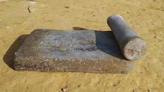 A traditional stone grinder set on a sandy surface, featuring a flat rectangular base slab and a cylindrical rolling stone. The surface of the stone is worn and slightly discolored, suggesting frequent use. Sunlight casts shadows, enhancing the texture of the grinder.