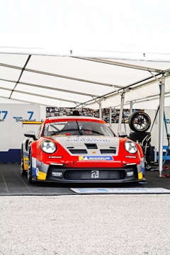 A race car with a prominent Porsche logo is positioned under a white tent. The car is in red, white, and yellow livery and features multiple sponsor decals. The area around the car is part of a pit garage, and a spare tire is visible to the right.
