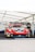 A race car with a prominent Porsche logo is positioned under a white tent. The car is in red, white, and yellow livery and features multiple sponsor decals. The area around the car is part of a pit garage, and a spare tire is visible to the right.