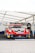 A race car with a prominent Porsche logo is positioned under a white tent. The car is in red, white, and yellow livery and features multiple sponsor decals. The area around the car is part of a pit garage, and a spare tire is visible to the right.