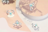 Several silver rings featuring opalescent gemstones are elegantly arranged on a soft, pastel surface. One of the rings is placed atop a small seashell, adding a delicate touch to the setting. Shadows from nearby foliage create interesting patterns on the background.