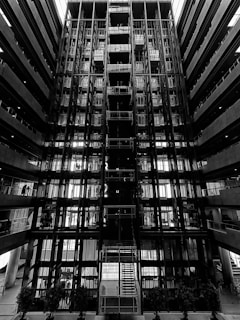 A symmetrical and striking architectural interior featuring multiple levels with glass and metal components. The central staircase is illuminated by natural light from above. The environment conveys an industrial and modern design with linear patterns.
