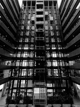 A symmetrical and striking architectural interior featuring multiple levels with glass and metal components. The central staircase is illuminated by natural light from above. The environment conveys an industrial and modern design with linear patterns.