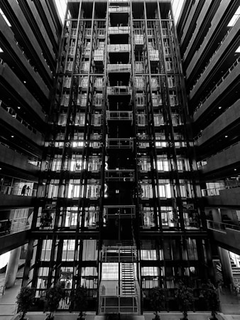 A symmetrical and striking architectural interior featuring multiple levels with glass and metal components. The central staircase is illuminated by natural light from above. The environment conveys an industrial and modern design with linear patterns.