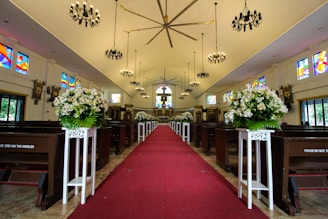 An ornate, well-lit church interior with a red carpet aisle running down the center, flanked by white floral arrangements on pedestals. The space is adorned with stained glass windows and dark chandeliers hanging from the ceiling. Wooden pews line the sides, and the altar is visible in the background.