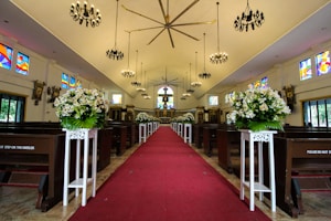 An ornate, well-lit church interior with a red carpet aisle running down the center, flanked by white floral arrangements on pedestals. The space is adorned with stained glass windows and dark chandeliers hanging from the ceiling. Wooden pews line the sides, and the altar is visible in the background.