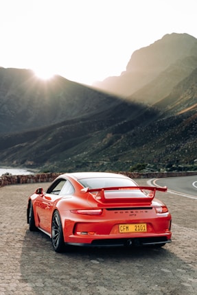 A sleek red sports car is parked on a scenic mountain road. The car, with its aerodynamic design and visible spoiler, stands out against the backdrop of rugged mountains. The sun is low in the sky, casting a soft glow and creating long shadows on the ground.