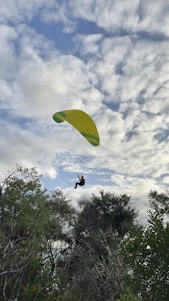 A paraglider is soaring through a partly cloudy sky, with lush green trees visible below. The yellow and green parachute contrasts with the white and blue clouds, giving a sense of adventure and freedom.