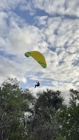 A paraglider is soaring through a partly cloudy sky, with lush green trees visible below. The yellow and green parachute contrasts with the white and blue clouds, giving a sense of adventure and freedom.