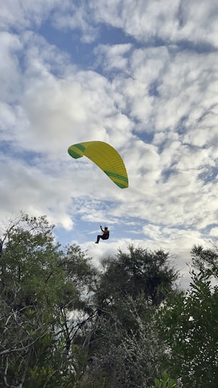 A paraglider is soaring through a partly cloudy sky, with lush green trees visible below. The yellow and green parachute contrasts with the white and blue clouds, giving a sense of adventure and freedom.