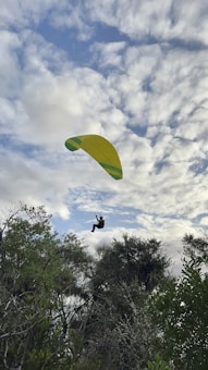A paraglider is soaring through a partly cloudy sky, with lush green trees visible below. The yellow and green parachute contrasts with the white and blue clouds, giving a sense of adventure and freedom.