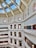 A grand, multi-story library interior with ornate architecture and a large, arched glass ceiling. The library features rows of bookshelves filled with books, and decorative columns and arches. Natural light streams in through the skylights, illuminating the detailed molding and lamps that hang along the railings.