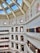 A grand, multi-story library interior with ornate architecture and a large, arched glass ceiling. The library features rows of bookshelves filled with books, and decorative columns and arches. Natural light streams in through the skylights, illuminating the detailed molding and lamps that hang along the railings.