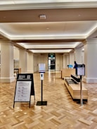 A spacious interior of a public building with polished wooden flooring and a high ceiling. There are informational stands and a hand sanitizer dispenser in the foreground. A reception desk is visible further back, with a person standing behind it. There is an entrance or exit door in the background, flanked by large columns.