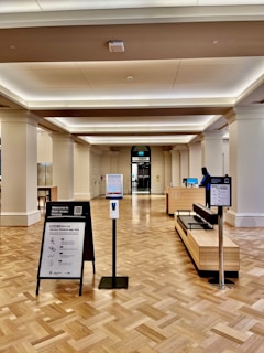 A spacious interior of a public building with polished wooden flooring and a high ceiling. There are informational stands and a hand sanitizer dispenser in the foreground. A reception desk is visible further back, with a person standing behind it. There is an entrance or exit door in the background, flanked by large columns.
