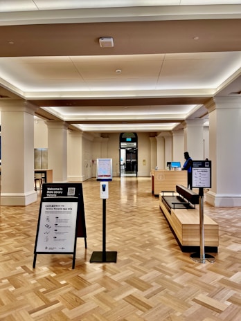 A spacious interior of a public building with polished wooden flooring and a high ceiling. There are informational stands and a hand sanitizer dispenser in the foreground. A reception desk is visible further back, with a person standing behind it. There is an entrance or exit door in the background, flanked by large columns.