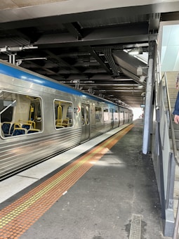 A sleek, silver train is stationed on the track under a roofed platform. The train has large windows and blue stripes running along the top. The platform features a tactile caution strip in orange and yellow. Overhead, there are visible metal beams and conduits, contributing to the industrial aesthetic. A partial view of a staircase and a handrail is visible on the right side.
