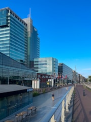 A modern urban scene featuring a row of glass-fronted high-rise buildings under a clear blue sky. The forefront has a pedestrian walkway with a few people walking, flanked by outdoor seating and a cafe area. Colorful flags are displayed along the path, and there are bicycles parked nearby.