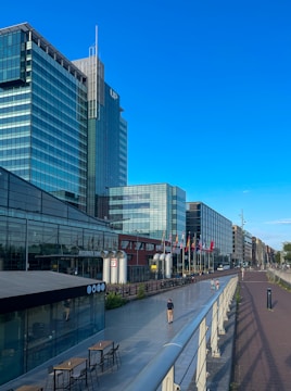 A modern urban scene featuring a row of glass-fronted high-rise buildings under a clear blue sky. The forefront has a pedestrian walkway with a few people walking, flanked by outdoor seating and a cafe area. Colorful flags are displayed along the path, and there are bicycles parked nearby.