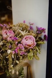 A bouquet of flowers featuring pink peonies with layers of soft petals, interspersed with small purple and white blooms. The arrangement includes lush green leaves and dried golden sprigs. The bouquet is set against a simple, neutral background, highlighting the vivid colors of the flowers.