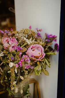 A bouquet of flowers featuring pink peonies with layers of soft petals, interspersed with small purple and white blooms. The arrangement includes lush green leaves and dried golden sprigs. The bouquet is set against a simple, neutral background, highlighting the vivid colors of the flowers.
