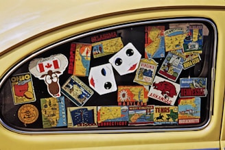 A car window filled with colorful stickers representing different U.S. states and Canada. The stickers display state names, maps, and iconic symbols, such as a maple leaf for Canada and a cartoon moose. The stickers are arranged haphazardly across the window, creating a collage effect.