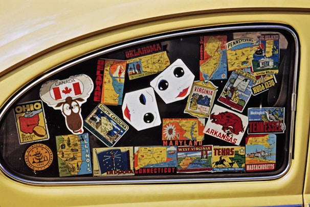 A car window filled with colorful stickers representing different U.S. states and Canada. The stickers display state names, maps, and iconic symbols, such as a maple leaf for Canada and a cartoon moose. The stickers are arranged haphazardly across the window, creating a collage effect.